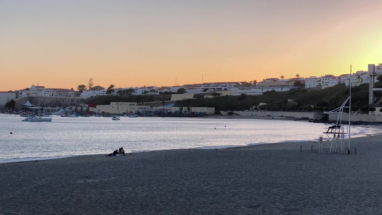 Villaggio costiero con una baia, barche e persone che si rilassano sulla spiaggia durante un bellissimo tramonto, offrendo temi di viaggio e lifestyle