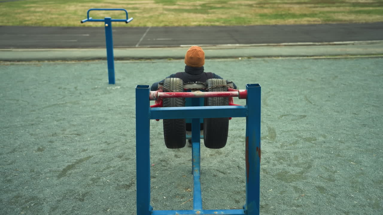 A coach in a brown beanie working out on an outdoor exercise machine with large tires, while a person in black clothing jogs on a nearby track adjacent to a grassy field