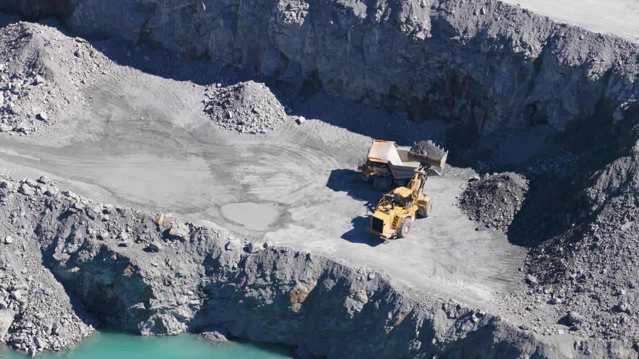 Telephoto drone captures trucks and excavator in active quarry. Bright daylight highlights industrial activity and rugged terrain