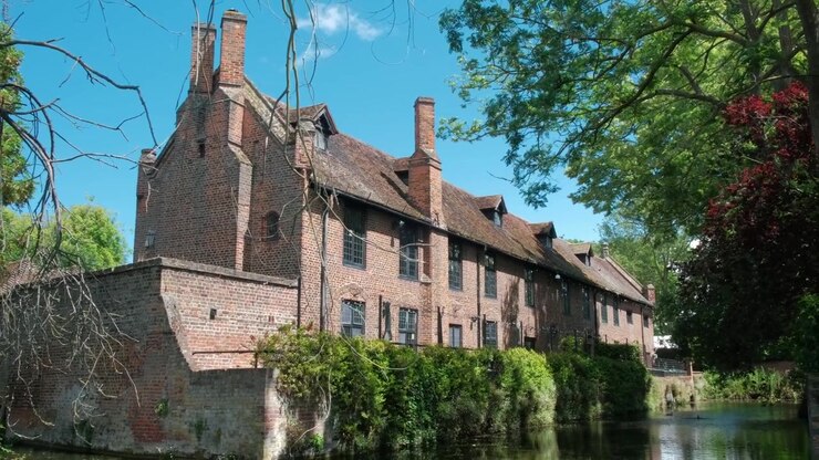 Vista tranquilla sull'acqua di un edificio storico in mattoni rossi a Well Hall Park, Eltham, con vegetazione lussureggiante, cieli azzurri e riflessi architettonici sullo stagno
