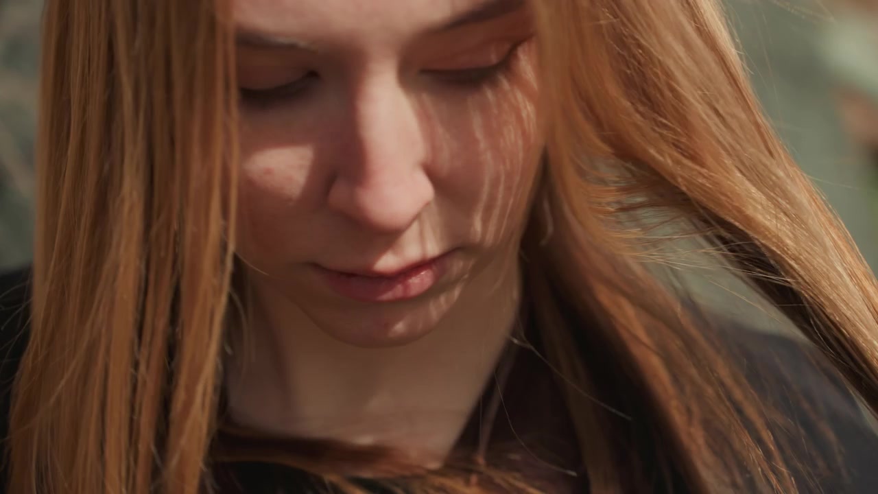 Close-up portrait of person with long reddish blonde hair gently gathering strands with hands while looking downward, showing subtle smile, fingerless glove, layered sleeves, warm outdoor light