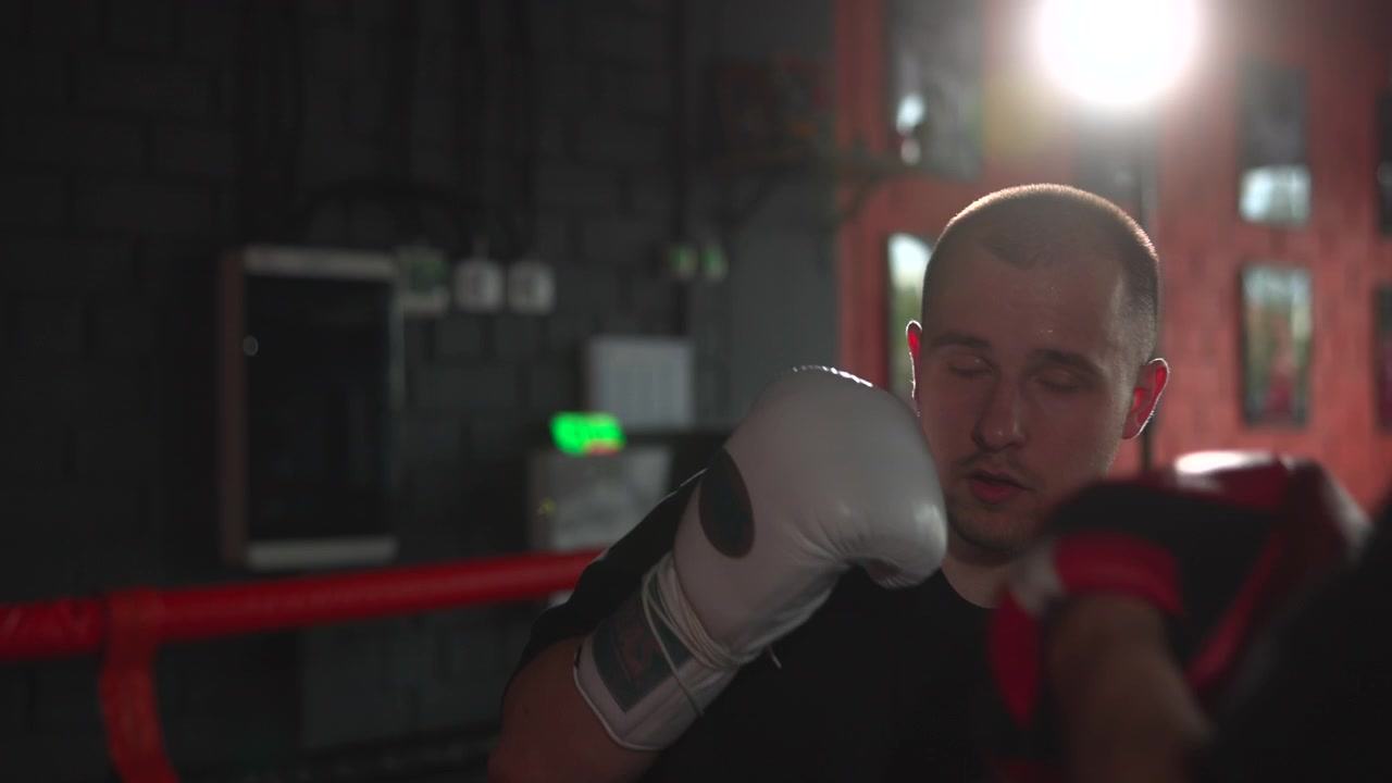 Young male boxer stands inside dim indoor ring with both white gloves raised in tight defensive guard, focused gaze, red ropes, brick wall backdrop, moody light, intense training atmosphere