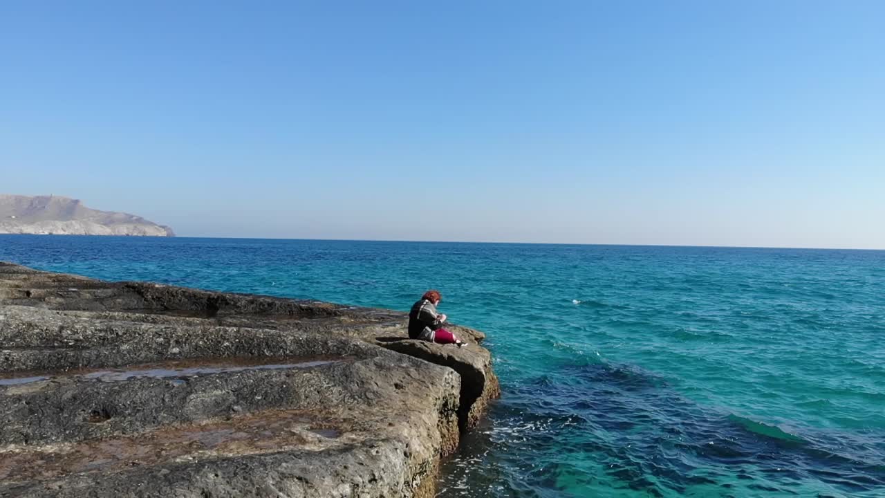 뒤에 아름 다운 푸른 물과 함께 책을 읽고 바위에 앉아 소녀의 공중 추적 샷