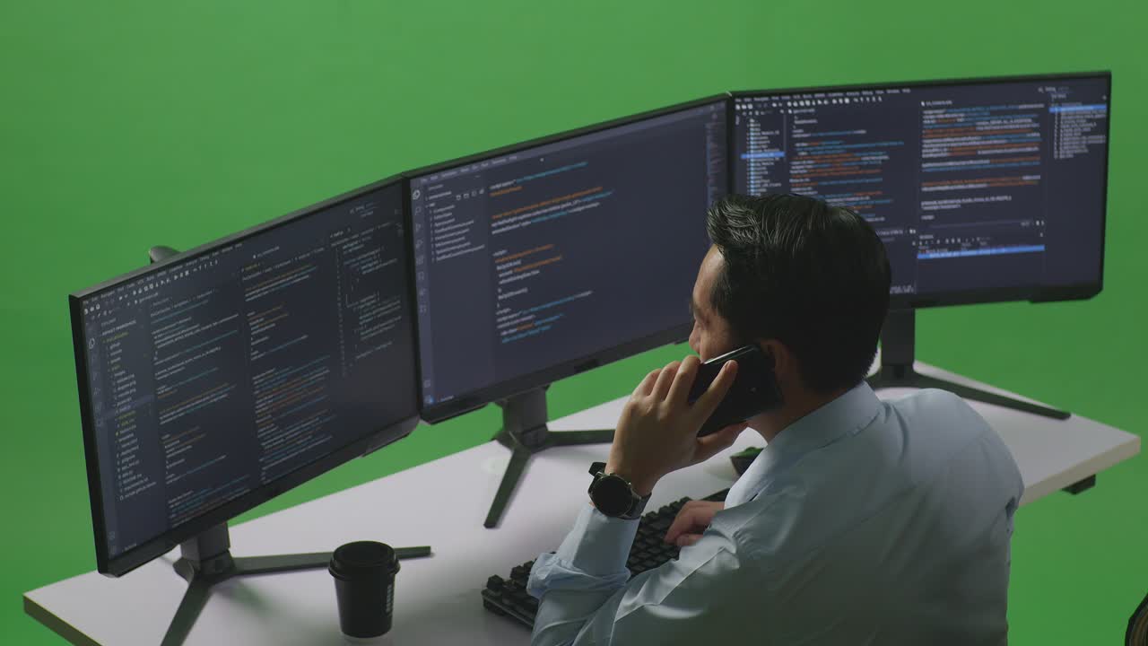 Side View Of Asian Man Developer Talking On Mobile Phone While Write Code With Multiple Computer Screens In Green Screen Studio