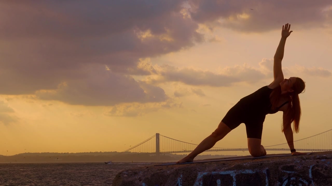 Donna che pratica la posizione yoga della plancia laterale in ginocchio all'aperto. Silhouette che estende il braccio verso l'alto su una sporgenza di cemento, Ponte di Verrazzano Narrows, porto di New York, tramonto