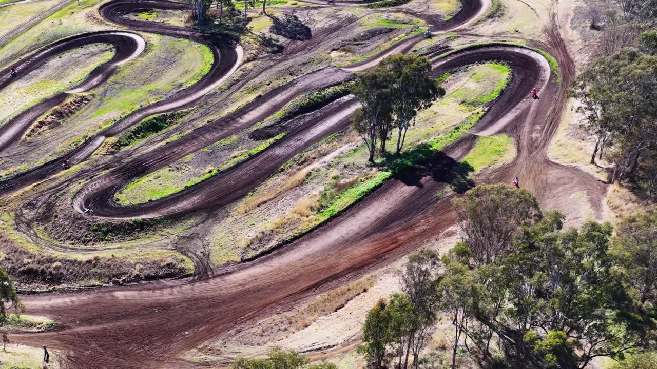 Drone smoothly flies above winding dirt motocross track, trees, and rural road in bright daylight