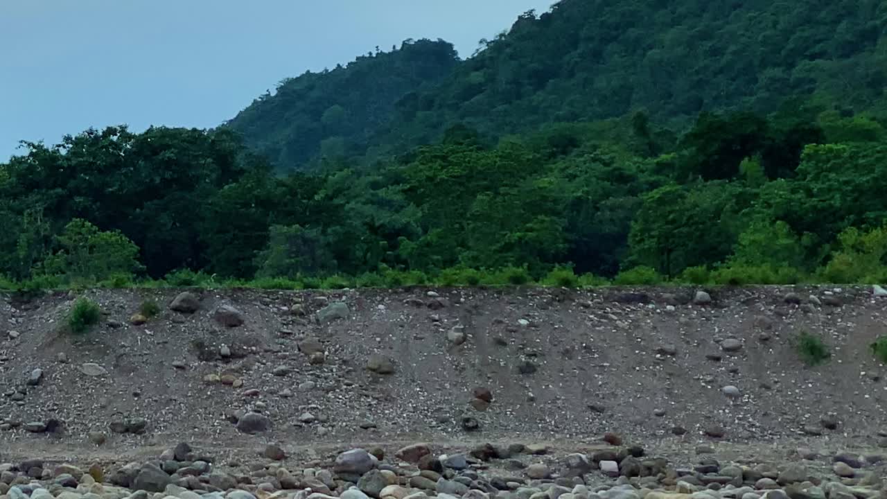 Aliran lebar berisi kerikil dengan bebatuan yang tersebar di bawah tanggul kerikil dan perbukitan berhutan yang rimbun di Bichnakandi, Sylhet, Bangladesh