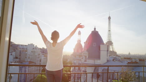 Joven-Turista-Feliz-Con-Los-Brazos-Levantados-Celebrando-Exitosas-Vacaciones-De-Viaje-Disfrutando-De-La-Libertad-Independiente-Relajándose-En-El-Balcón-Al-Atardecer-En-París,-Francia,-Mirando-La-Torre-Eiffel