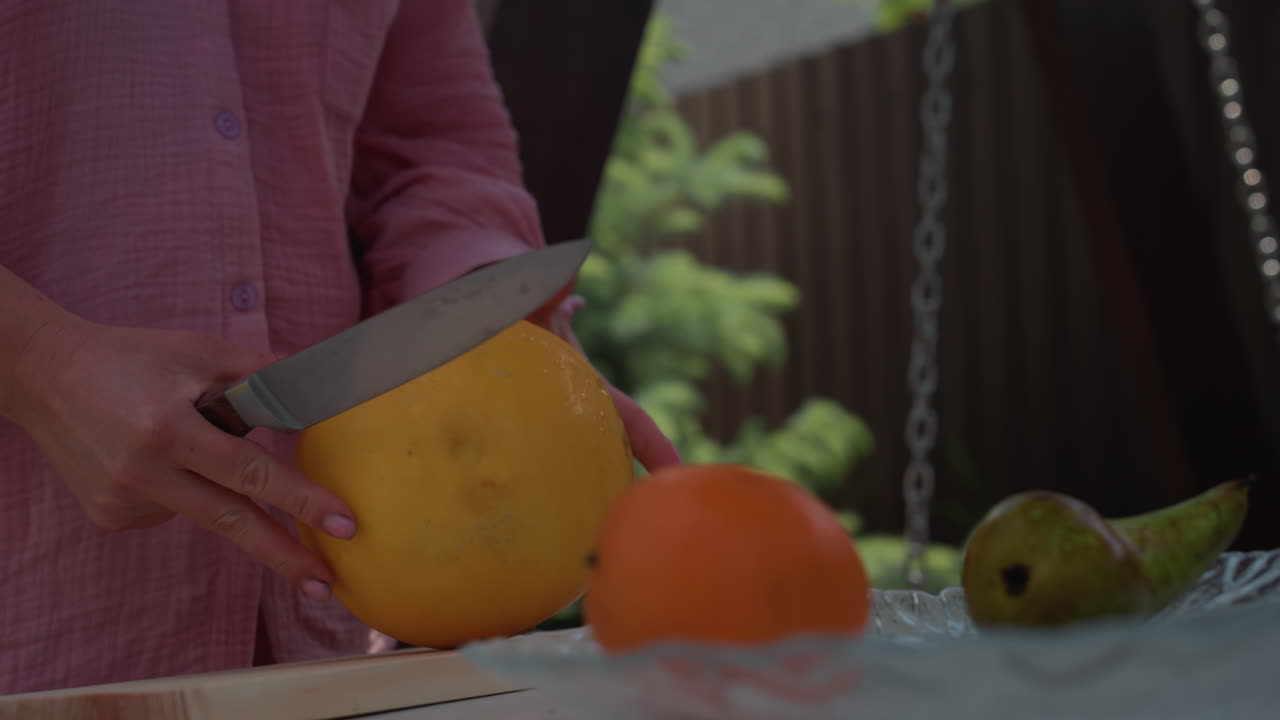 Woman Holding Melon And Knife At Table, Midshot Hands Slicing Round Yellow Melon Beside Tangerine And Pear, Outdoor Backyard Harvest Scene, Careful Preparation Under Sunlight, Rustic Homegrown Produce