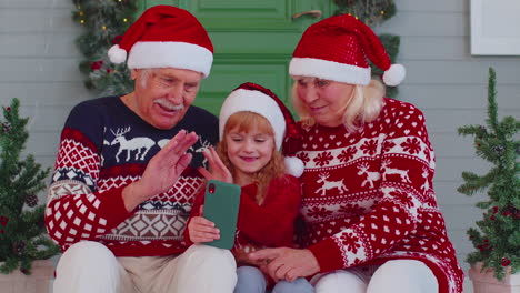 Smiling-grandparents-and-granddaughter-waving-during-video-call-on-smartphone-at-Christmas-reunion