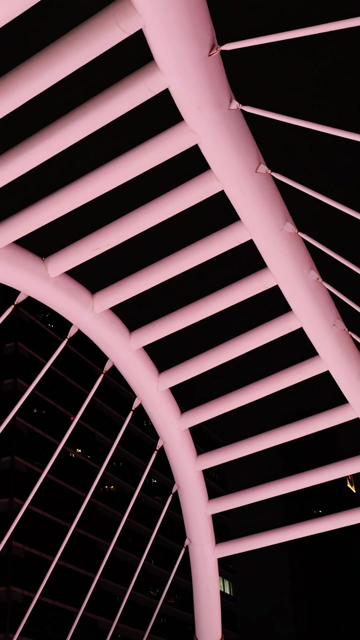 A câmera se inclina para cima, revelando a arquitetura moderna e iluminada de uma ponte contra o horizonte noturno da cidade, com iluminação dramática.
