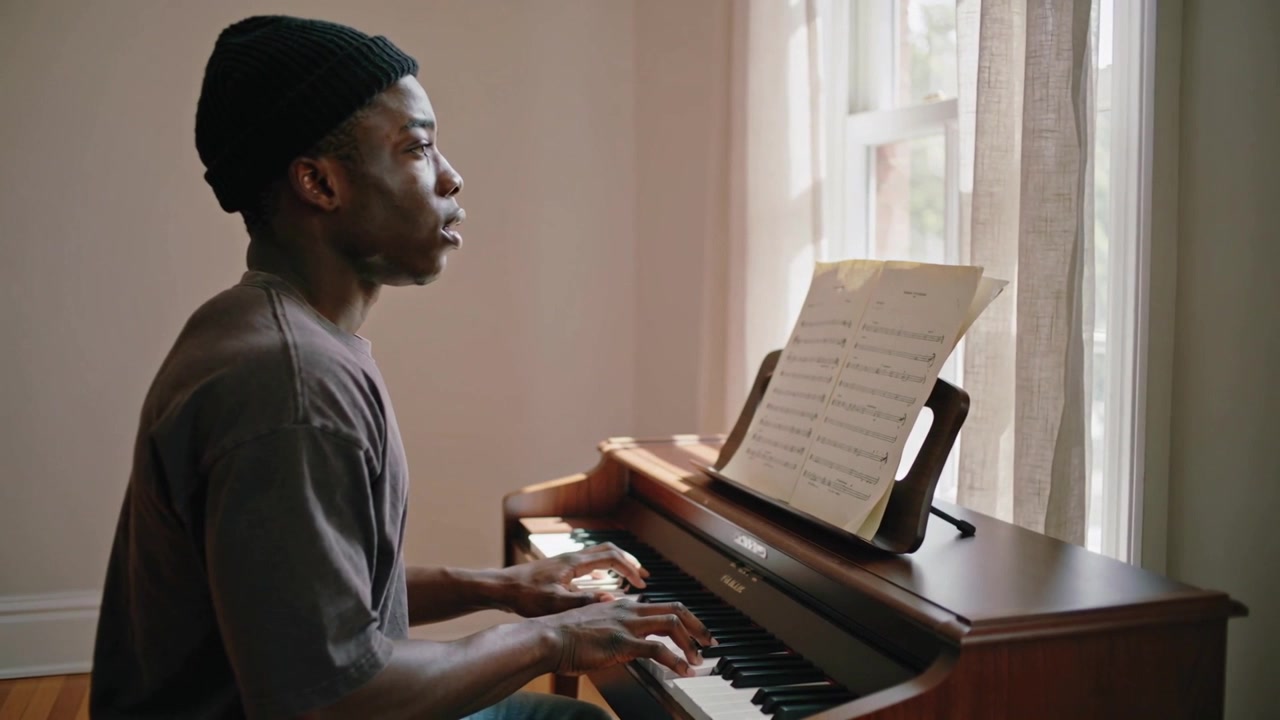 Reading sheet music, man in beanie and dark tee practicing digital piano at home, copy space