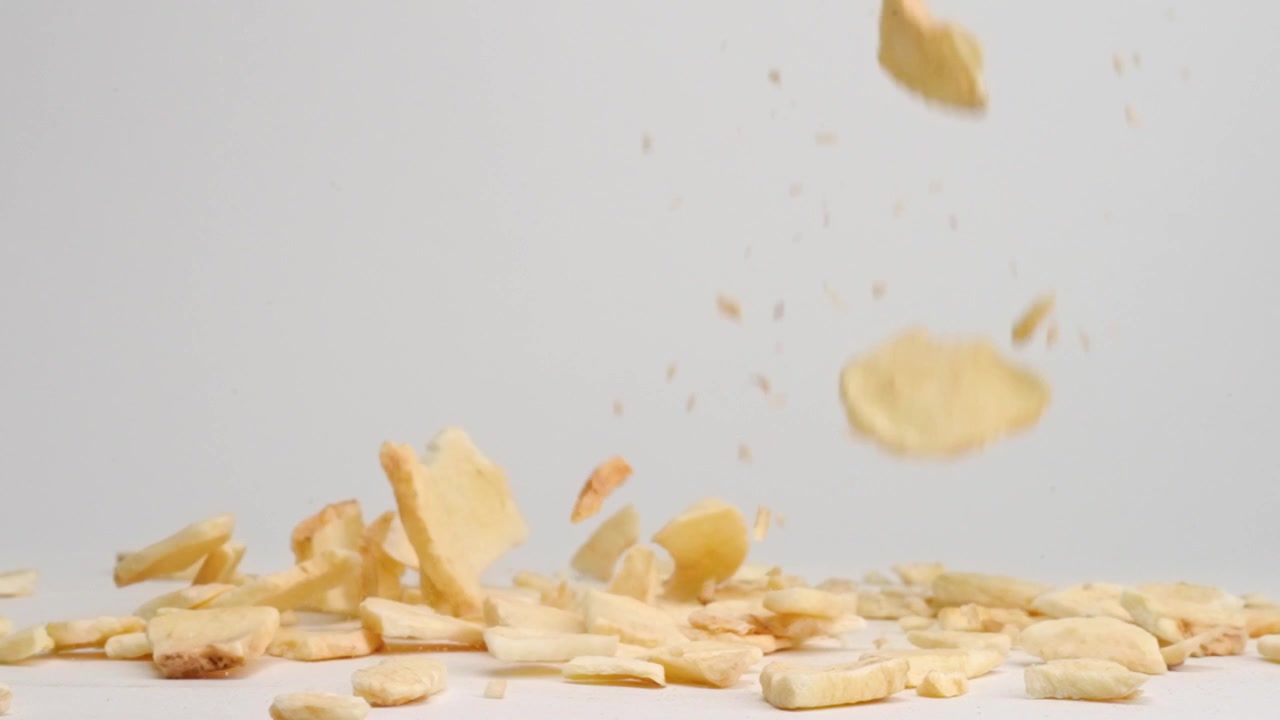 Pedaços de maçã desidratada caindo sobre uma mesa branca, quicando e formando uma pilha em câmara lenta