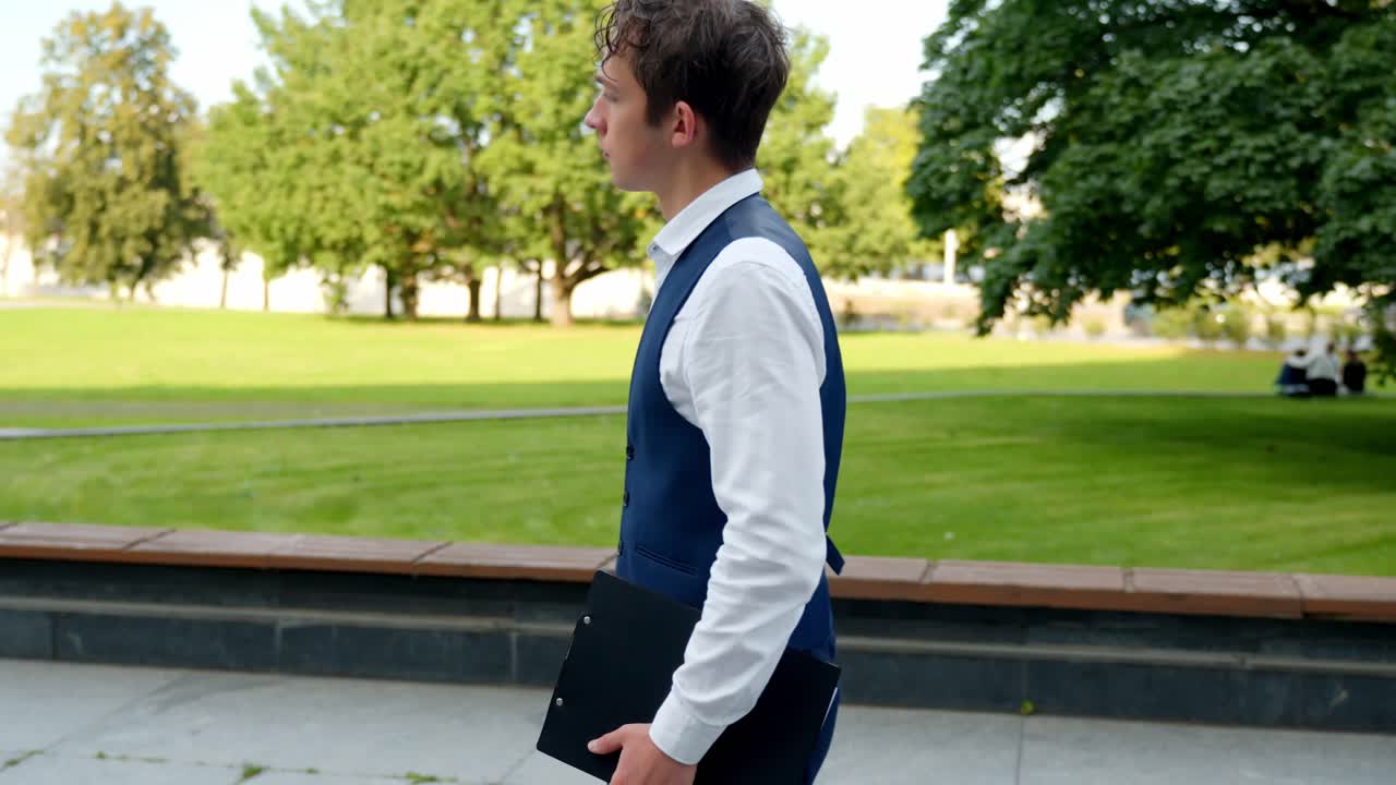 Young financial analyst business man walking confident in an outdoor park holding a document folder, representing success, career, and a professional lifestyle, slow motion shot