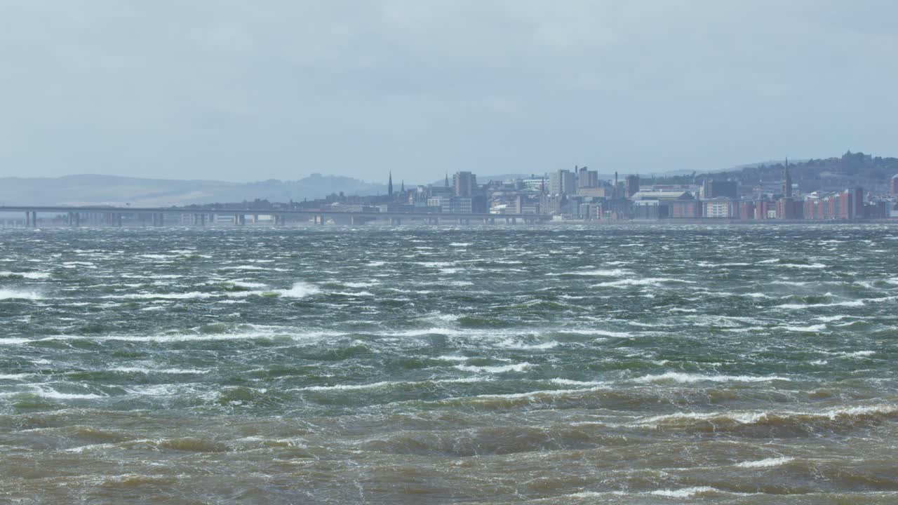 Choppy sea waves approach the beach with cityscape and hills in the background