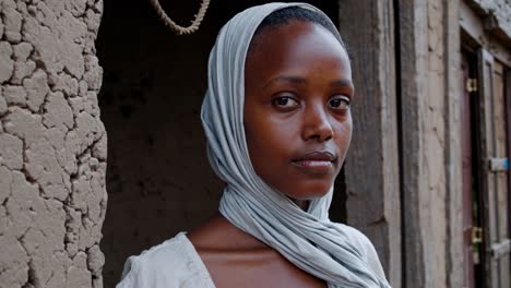 young african woman wearing traditional white dress and headscarf standing confidently in mud brick house doorway, embodying rural lifestyle and cultural resilience with serene, dignified presence