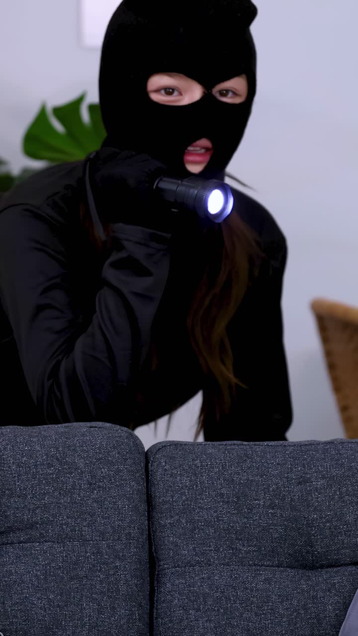 A masked intruder cautiously navigates a dark living room using a flashlight during a burglary