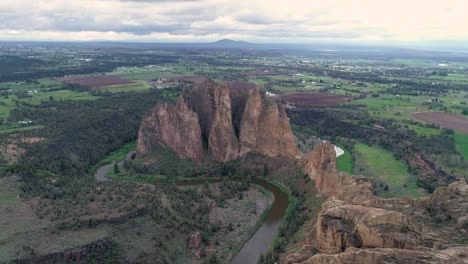 Una-Toma-Inversa-De-Un-Dron-En-Movimiento-Lento-De-Smith-Rock,-Oregón,-Un-Río-Que-Fluye-A-Su-Lado-Y-Las-Tierras-De-Cultivo-Circundantes.