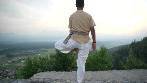 indian man doing hatha yoga standing at the edge of a stone castle wall in the morning sun at sunrise overlooking the valley bellow with fields and woods