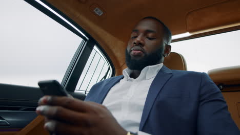 Closeup-guy-typing-mobile-phone-in-car.-Afro-businessman-chatting-smartphone