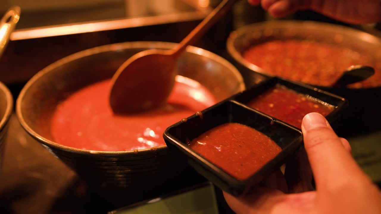 Hand ladles warm red sauce into black bowl under soft lighting at buffet station