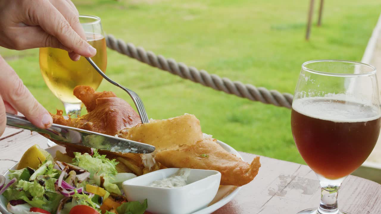 A person slices into battered fish with a fork and knife beside salad, tartar sauce, and beer on a sunlit outdoor table