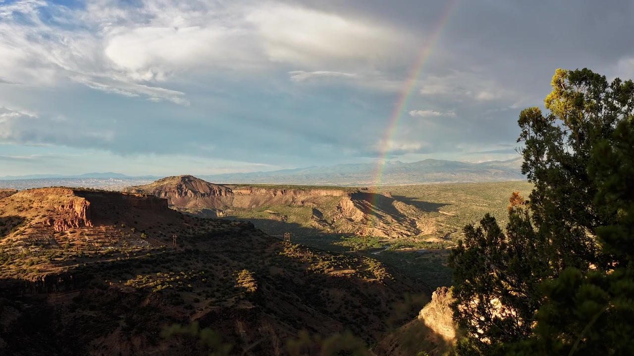 4K stabil timelapse av en stor frodig canyon. En lys regnbuebue forsvinner sakte over en dyp steinete dal og grønne trær mens tunge grå regnskyer og lette skygger driver over den vide himmelen