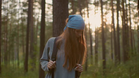 young girl in serene forest holding backpack straps, looking down contemplatively with sunlight softly filtering through trees behind her, creating a warm glow