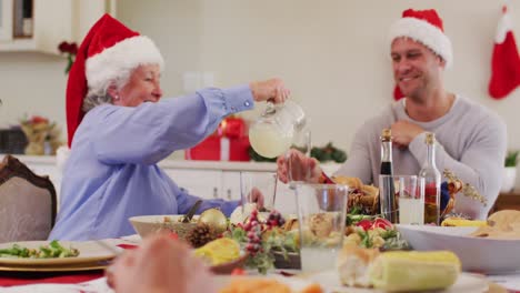 Kaukasische-Seniorin-Mit-Weihnachtsmütze-Gießt-Getränk-In-Ein-Glas-Ihres-Sohnes,-Während-Sie-Am-Esstisch-Sitzt