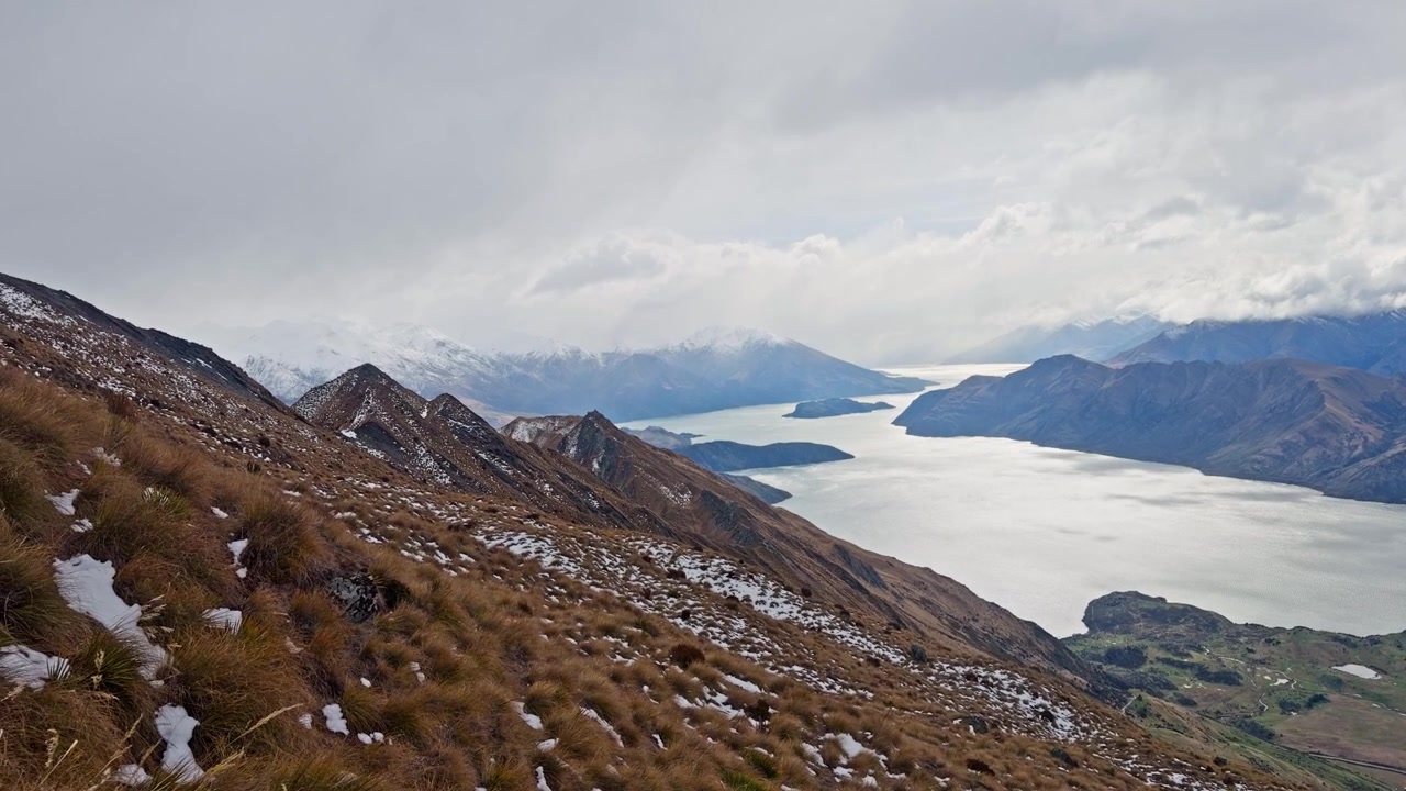 Alpine hiking trail with snow patches overlooking Lake Wanaka and distant mountains