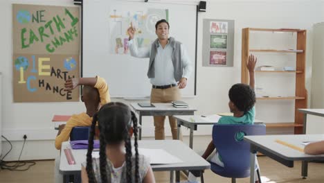 happy diverse male teacher teaching schoolchildren raising hands in classroom at elementary school