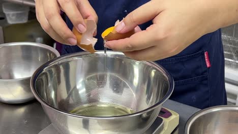 Sweet-dessert-making-in-progress,-pastry-chef-cracking-egg,-separating-egg-white-and-egg-yolk-into-different-stainless-steel-mixing-bowl-using-the-egg-shells,-commercial-restaurant-setting