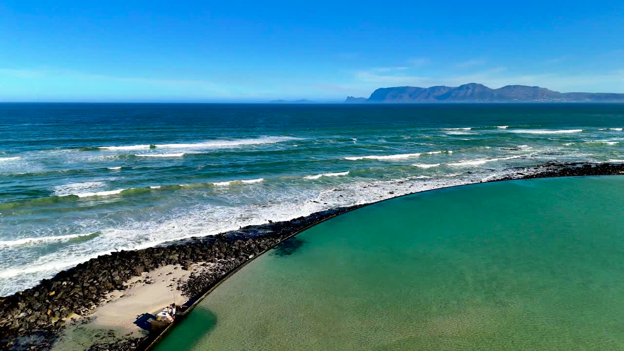 Drone ocean view from the edge of a tidal pool as waves break over rocks and a mountain range creates a scenic backdrop