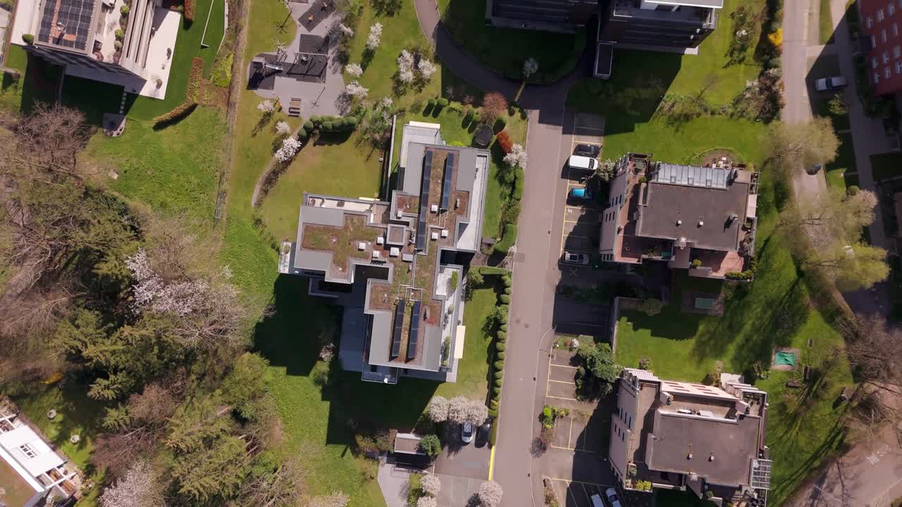 Solar panel on roof of residential apartments producing renewable energy on sunny day. Swiss small town with cars on street in alps. Green grass and lawn in garden. Aerial top down flyover.