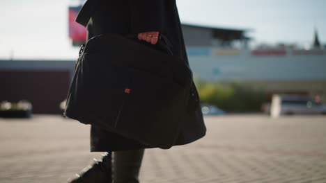 close-up of person carrying black bag with hand adorned with rings, walking outdoors in sunlight, dressed in black attire, background features blurred modern building and digital display