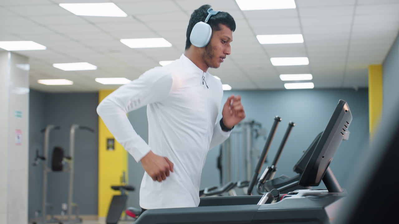 side view of young man running on treadmill in modern gym center with bright ceiling lights wearing headphones white top focused expression sleek equipment reflective mirror wall