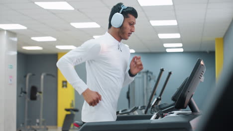 side view of young man running on treadmill in modern gym center with bright ceiling lights wearing headphones white top focused expression sleek equipment reflective mirror wall