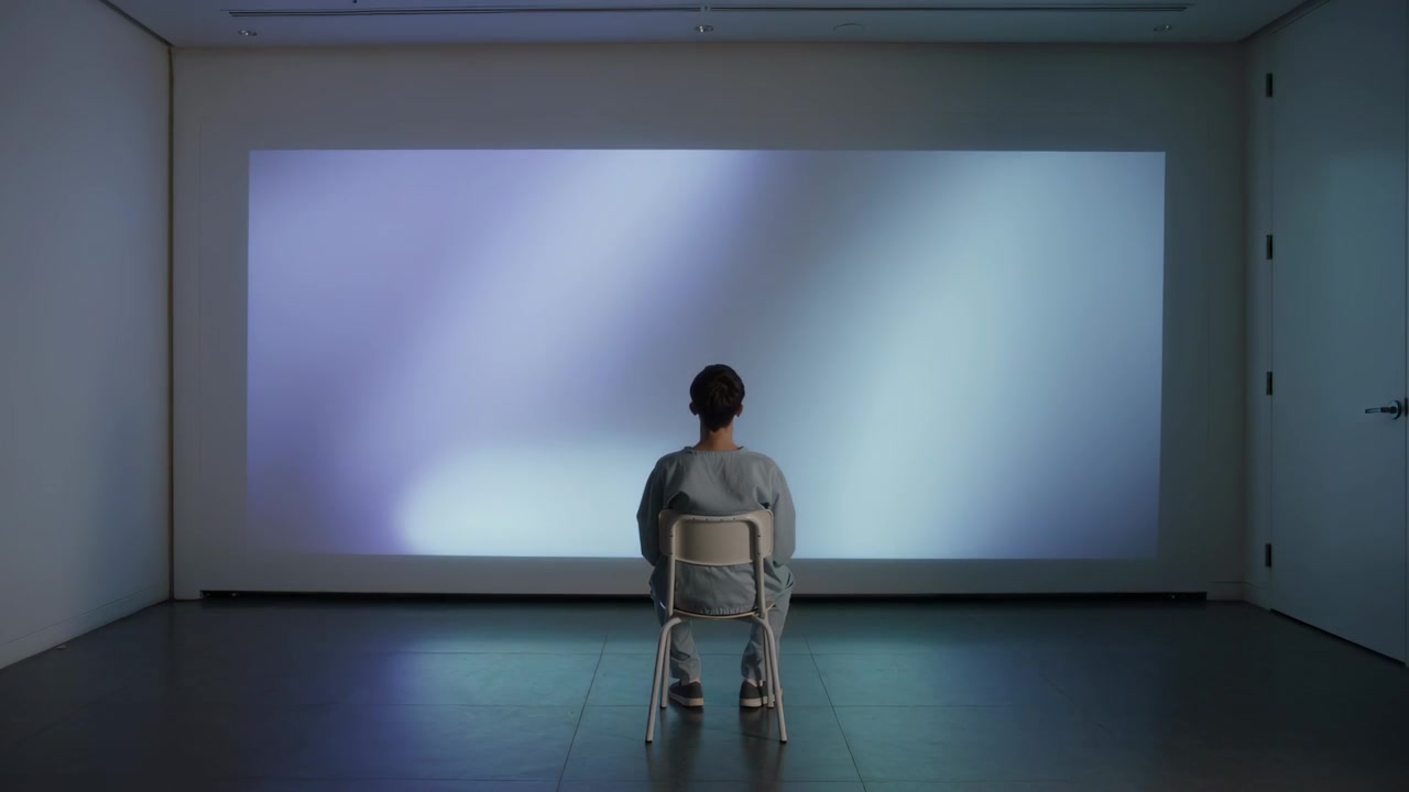Entering man taking metal folding chair, sitting in gallery watching blue projection, wearing gray