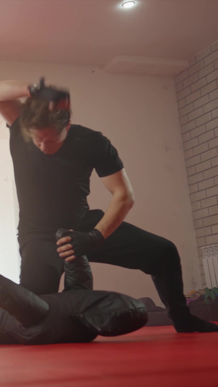 Young man practices martial arts techniques on red mat indoors. Man wears black clothing including gloves as man maintains. Setting features white brick walls with martial
