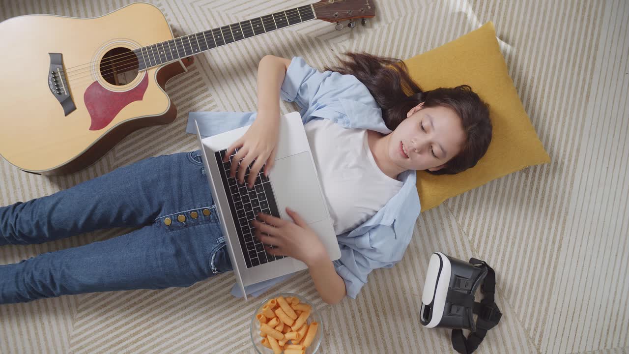 vista superior de una adolescente asiática con bocadillos escribiendo en una computadora portátil mientras yacía en la alfombra en el suelo de casa