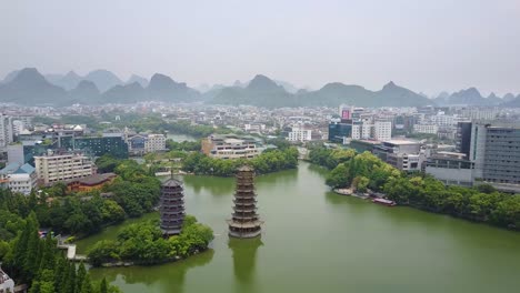 Aerial-Shot-of-the-Guilin-Sun-and-Moon-Pagodas-at-Lake-Shanhu-with-the-City-and-Jagged-Peak-in-the-Background,-Guangxi,-China