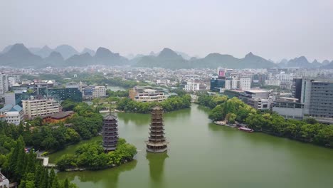 Toma-Aérea-De-Las-Pagodas-Del-Sol-Y-La-Luna-De-Guilin-En-El-Lago-Shanhu-Con-La-Ciudad-Y-El-Pico-Dentado-En-El-Fondo,-Guangxi,-China