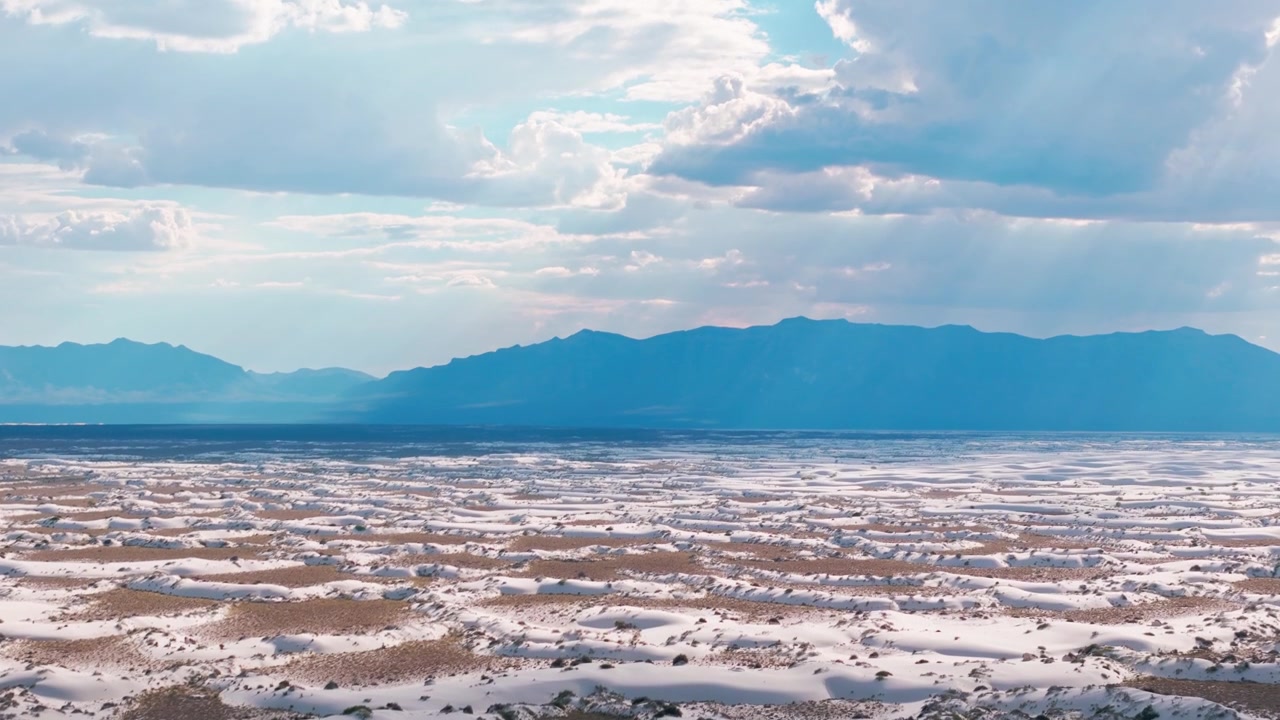 Panorama luftfoto i vidvinkel af glinsende gipsdyner i White Sands National Park, New Mexico, der viser den vidtstrakte tørre ørkenbund, naturlige teksturer og en fjern bjergkæde.