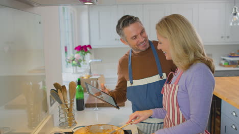 pareja madura en casa siguiendo la receta en la tableta digital en la cocina mientras preparan la comida juntos - filmado en cámara lenta