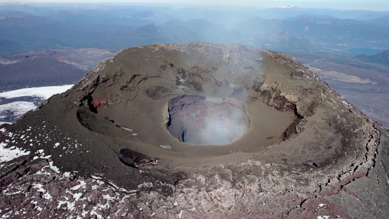 Ein atemberaubender Blick vom Gipfel eines Vulkans enthüllt einen großen Krater, umgeben von felsigem Gelände, mit einem ruhigen See in der Ferne vor einem klaren blauen Himmel.