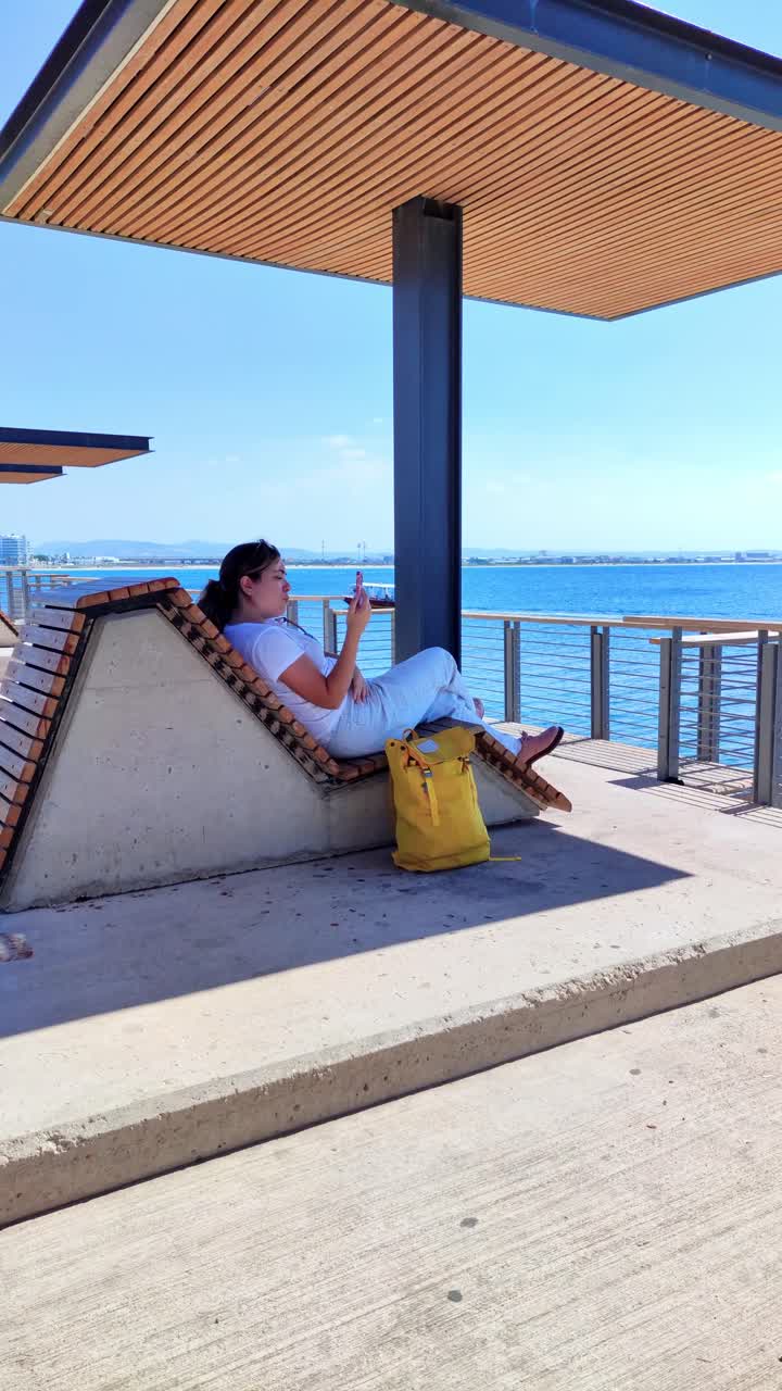 Giovane donna che si rilassa sotto un pergolato usando uno smartphone in riva al mare