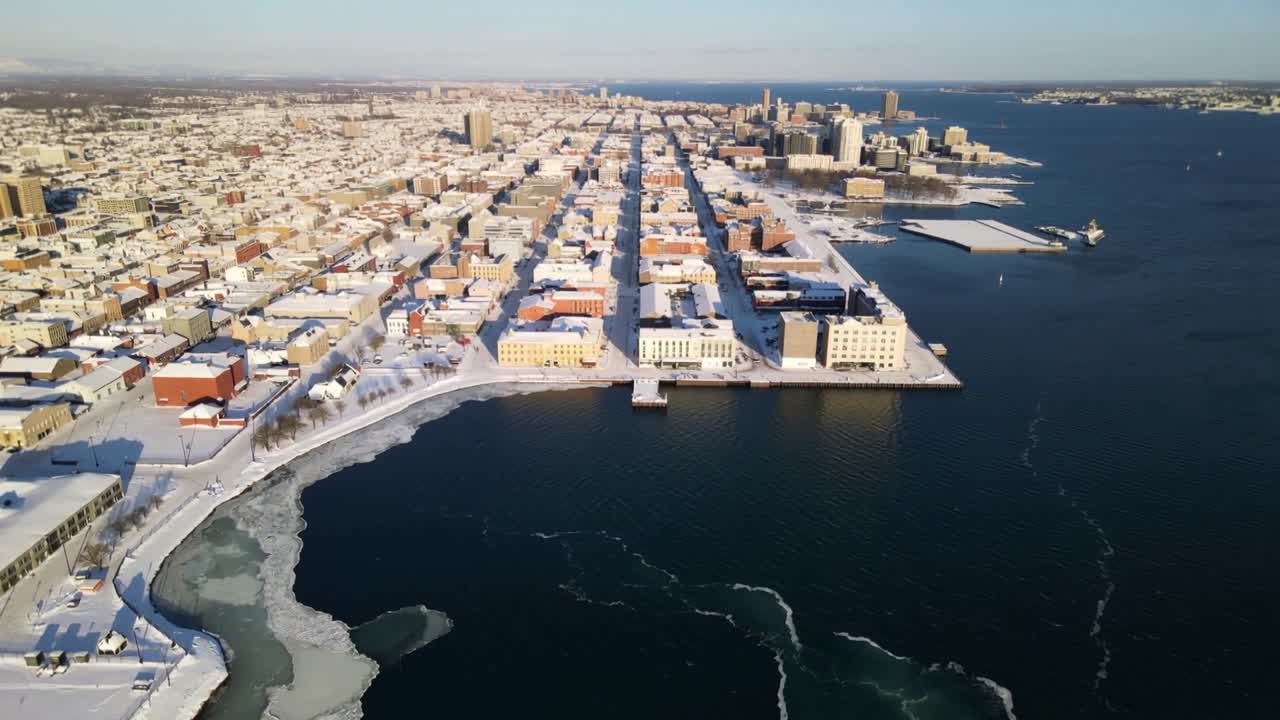 Fremrykkende drone avslører snødekt bygningsmasse ved vannkanten i havnen, nærmer seg brygge og isflak.