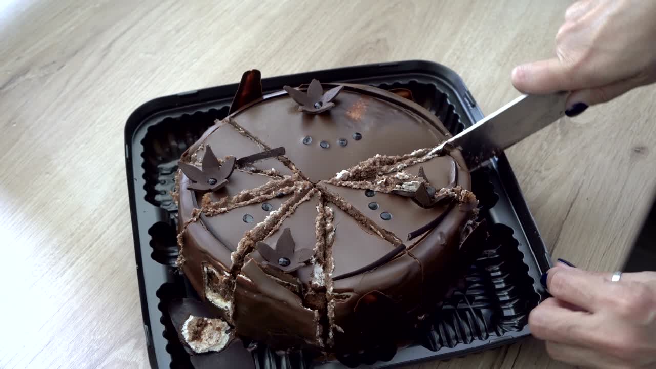 Donna taglia a pezzi grandi una torta al cioccolato decorata con fiori di cioccolato in cima. La torta è in una scatola regalo sul tavolo in cucina. Primo piano. Vista dall'alto. 4K