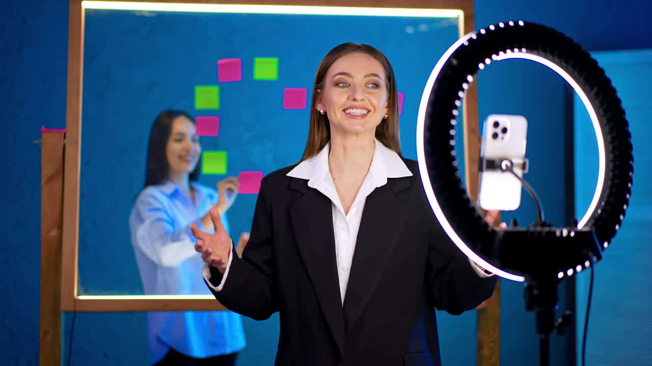 Caucasian smiling brunette wearing black jacket and white shirt stands in front of phone and ring light. Cheerful blogger turns to a colleague standing behind the glass board with stickers writing on it.