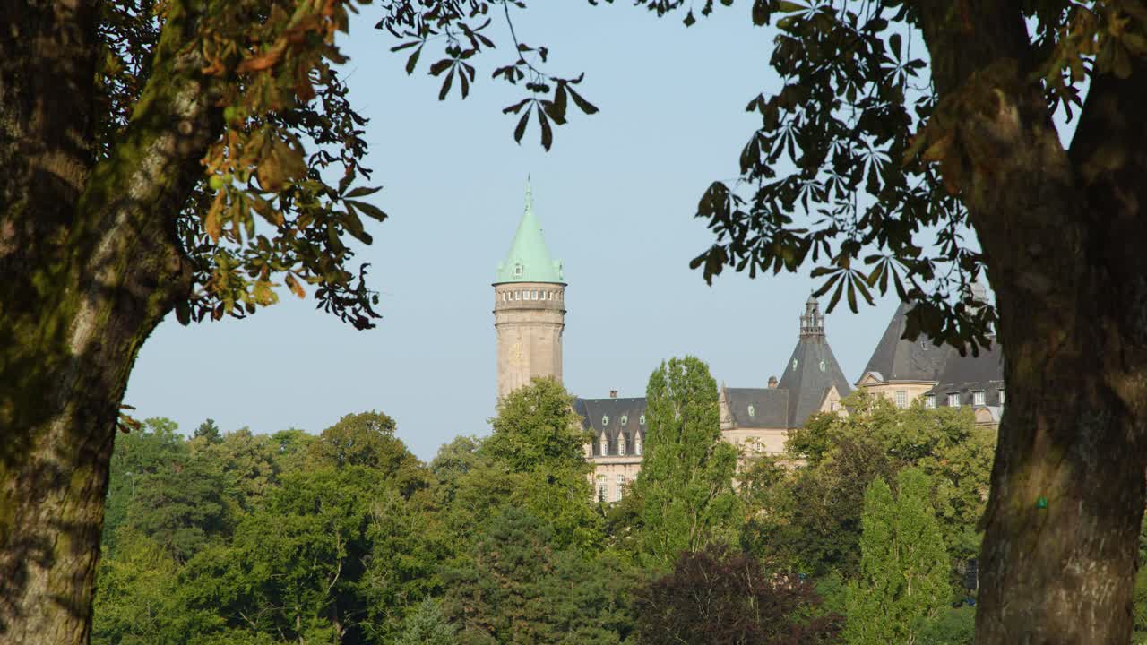 Static view of castle tower with green roof, framed by leafy trees, bright daylight, Luxembourg